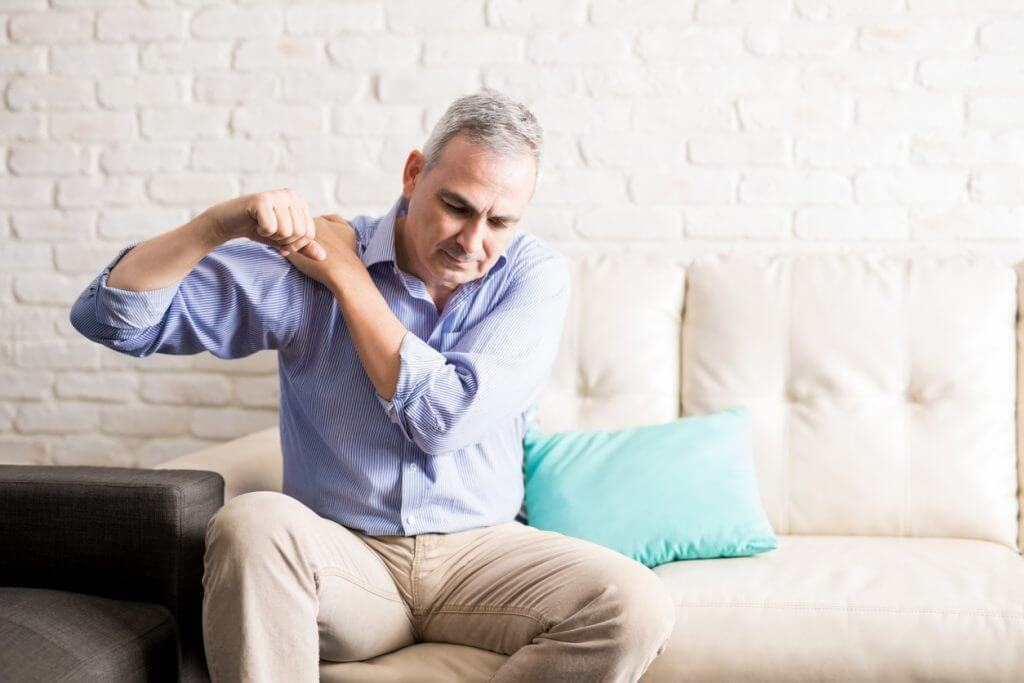 Middle aged man sitting on the sofa and holding painful shoulder with another hand.